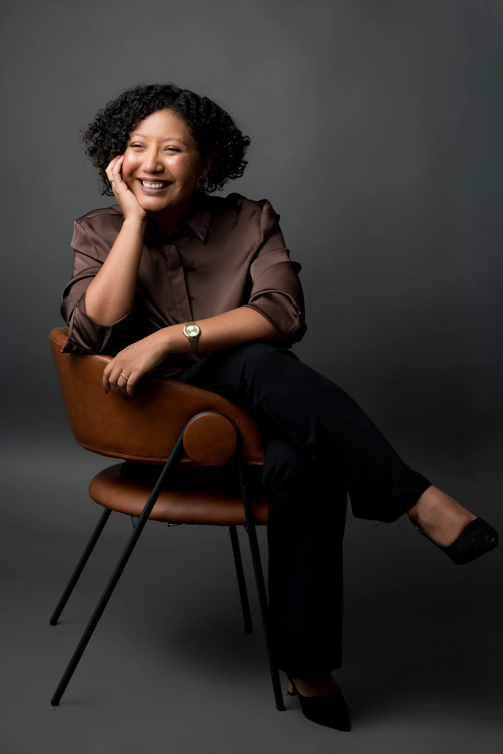 Margella Furtado smiling, seated in a leather chair, her hand on her cheek.